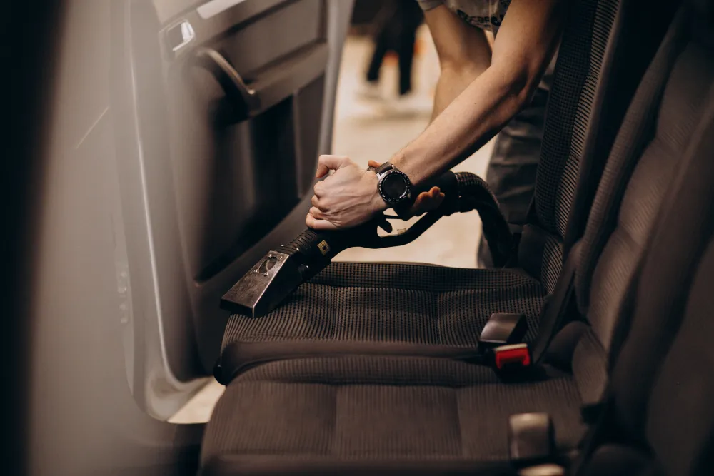 Person vacuuming the fabric seat of a car interior with a handheld vacuum cleaner.