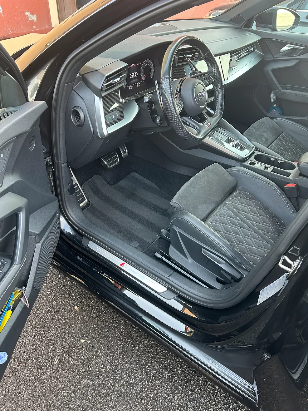Interior view of a black Audi car showing the driver's seat, steering wheel with Audi logo, digital dashboard, and door panel with controls.