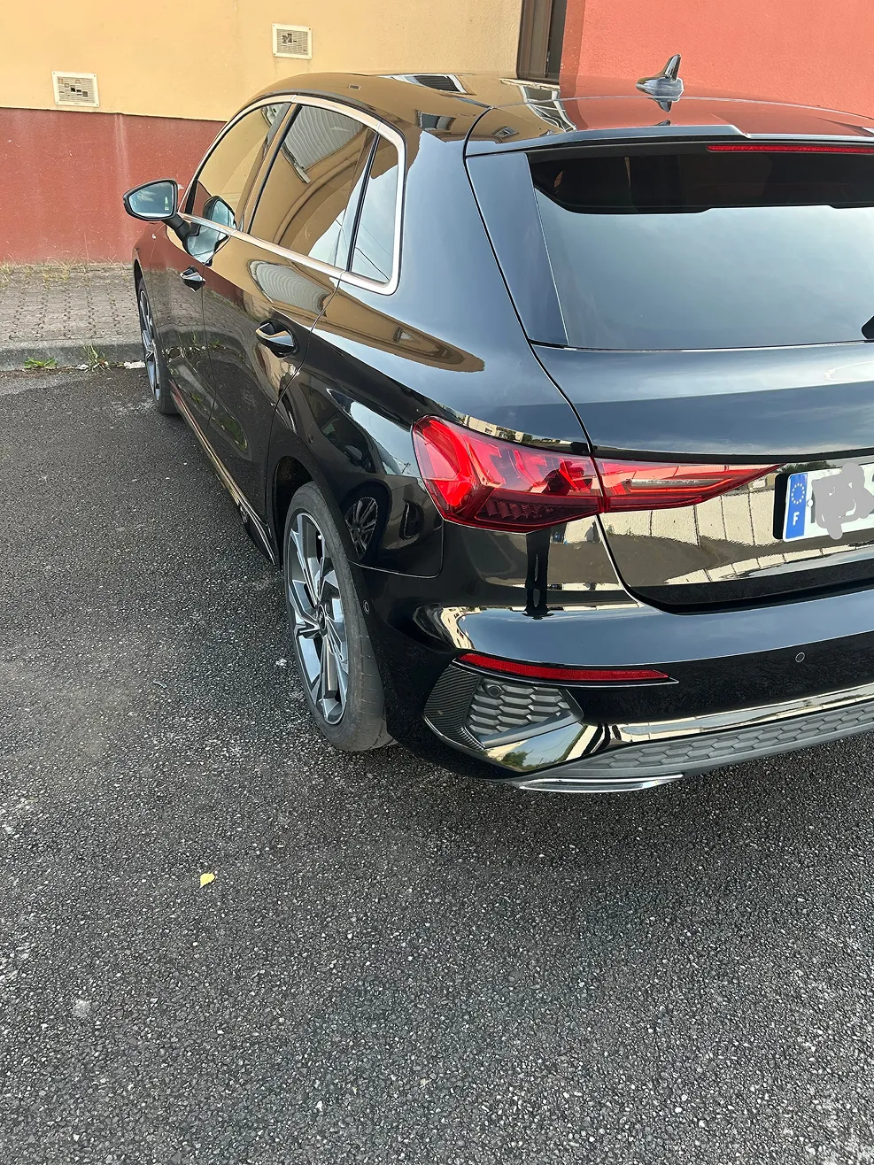 Rear-side view of a black hatchback car parked on asphalt near a red and yellow wall.