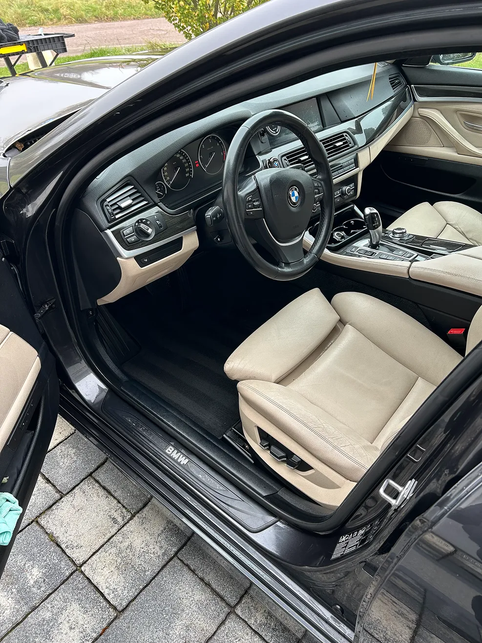 Interior view of a BMW car showing beige leather driver seat, steering wheel with BMW logo, dashboard, and center console gear shift.