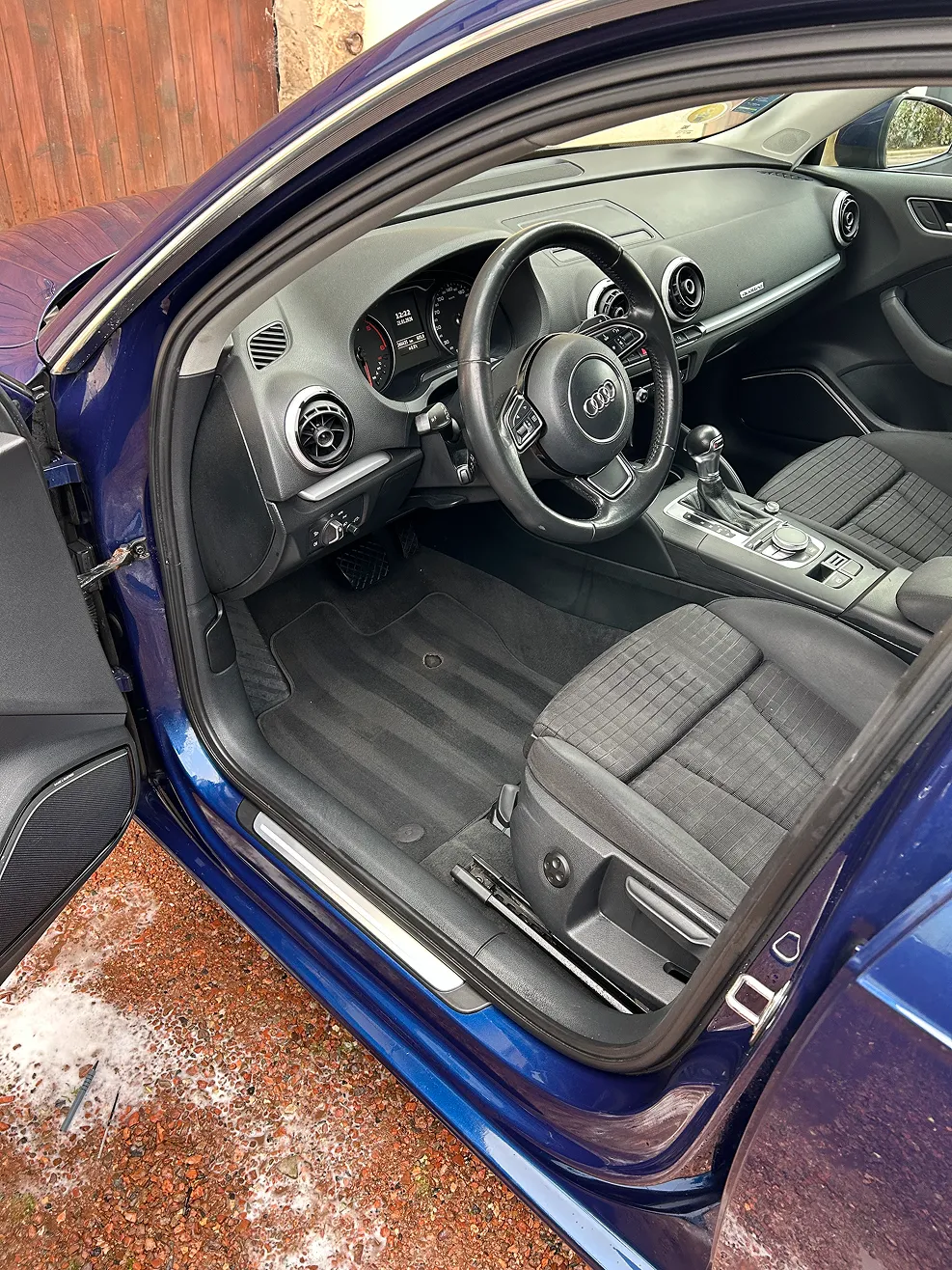 Interior view of a blue Audi car showing the driver's seat, steering wheel with Audi logo, dashboard, and center console with gear shift.