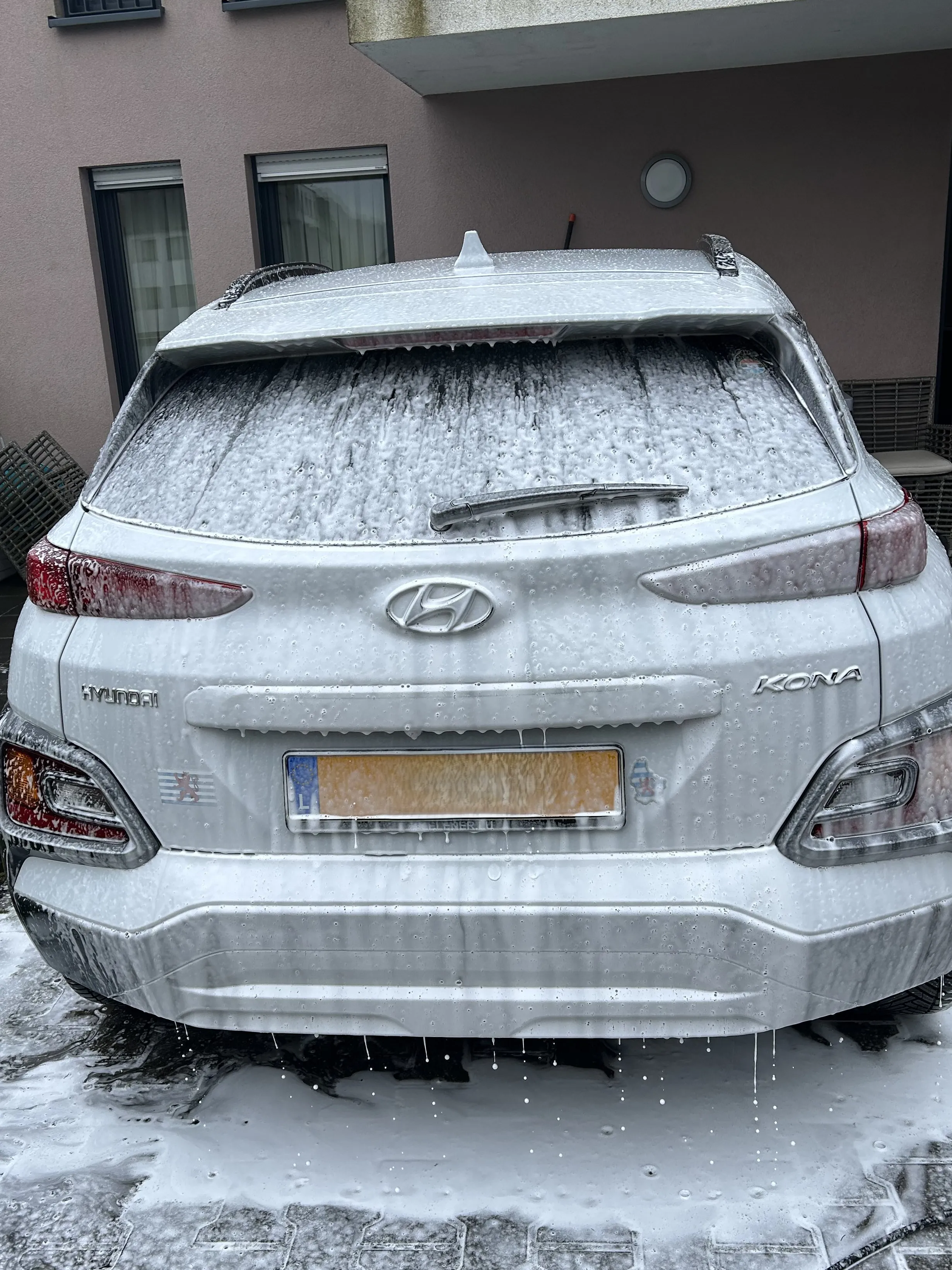 Rear view of a white Hyundai Kona covered in soap foam during a car wash.
