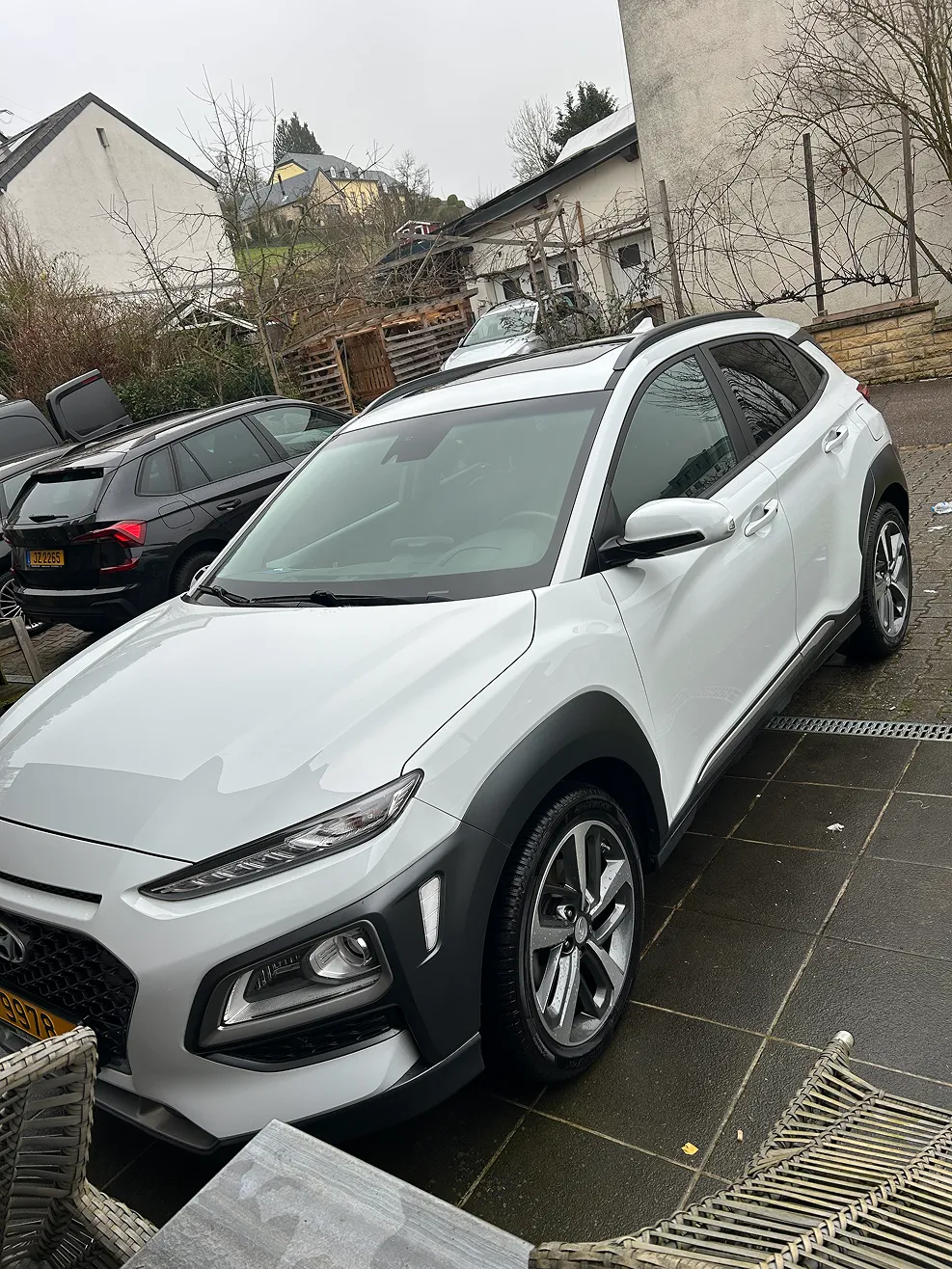 White Hyundai Kona parked on black tiled driveway with outdoor furniture nearby and houses in the background.