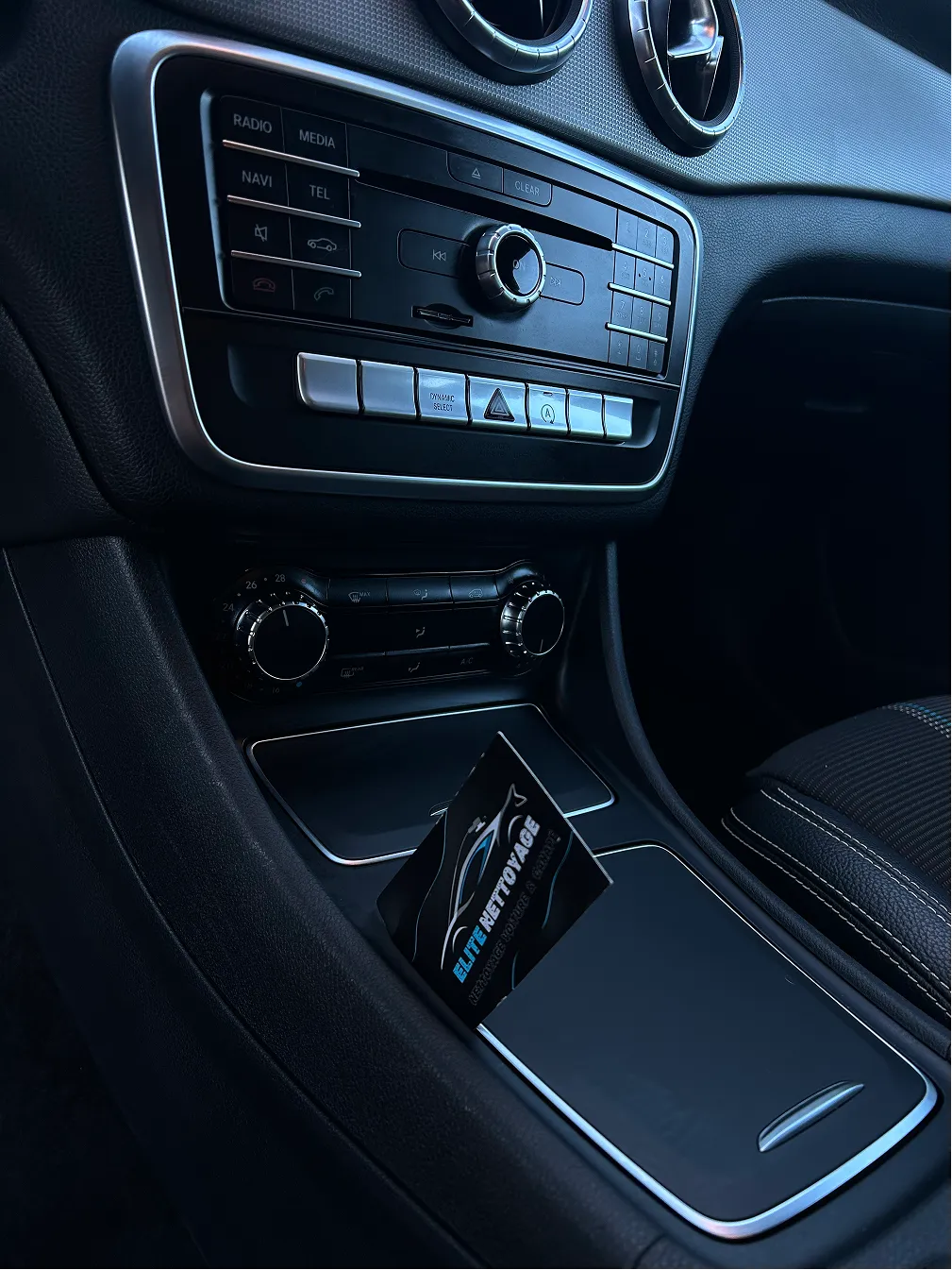 Center console of a car displaying media controls, air conditioning dials, and a card holder with an Elite Nettoyage business card.